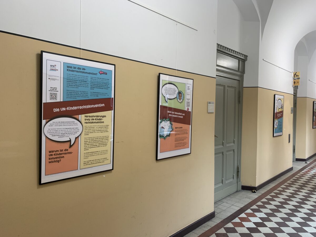 Schulflur mit gerahmten Informationsplakaten zur UN-Kinderrechtskonvention an einer beigefarbenen Wand.