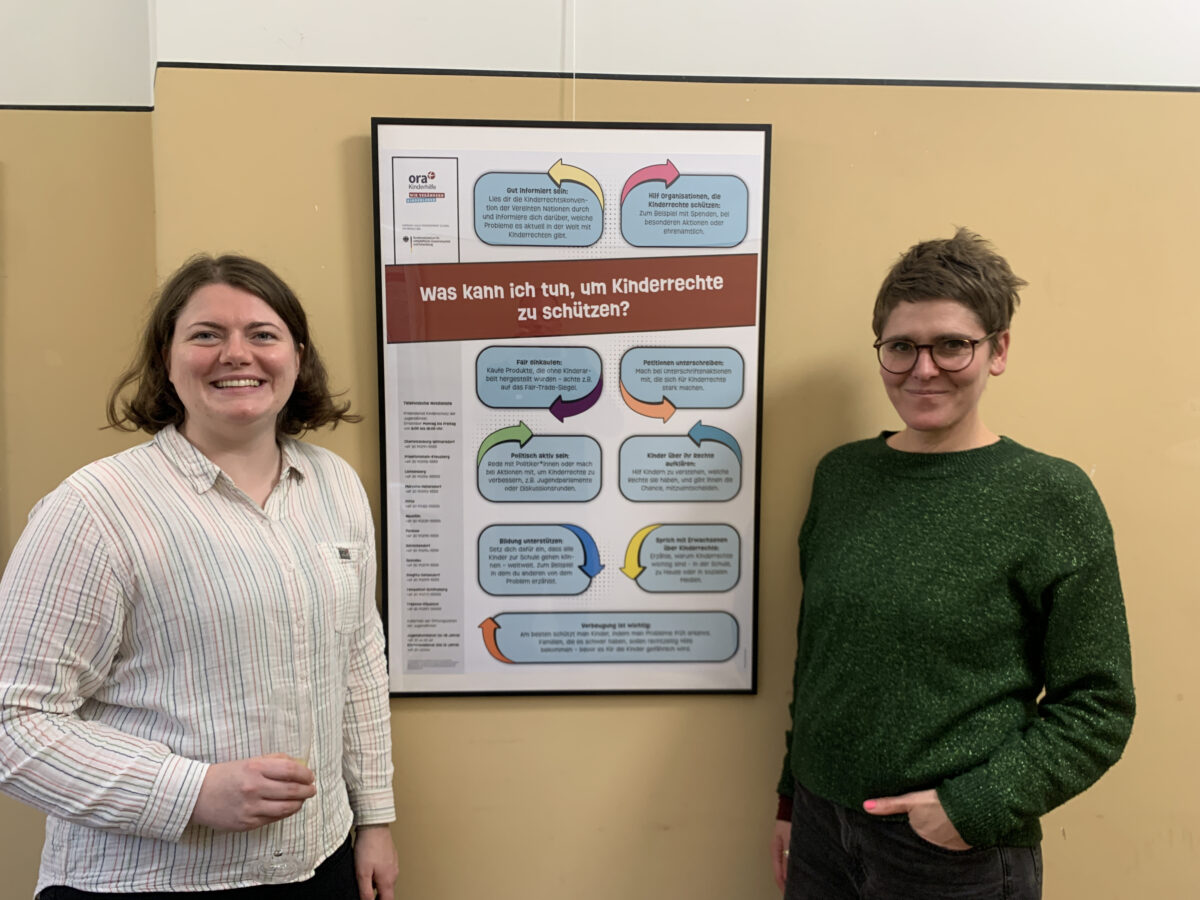 Zwei lächelnde Personen stehen neben einem Poster mit der Überschrift „Was kann ich tun, um Kinderrechte zu schützen?“.