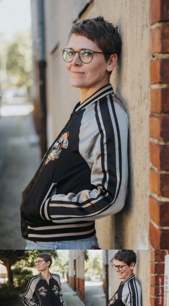 Porträt einer kurzhaarigen Frau mit Brille, die an einer Wand lehnt und eine schwarze-beige gestreifte Jacke trägt.